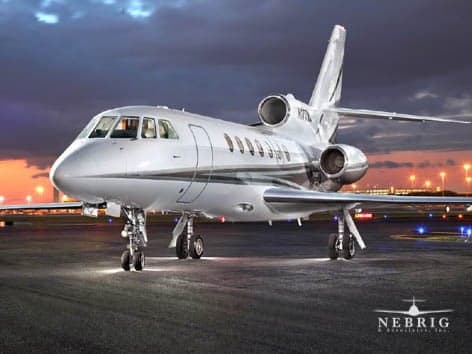 A silver Dassault Falcon business jet with dark grey stripes is parked on an tarmac at dusk, with ground lights illuminated and a sunset sky.
