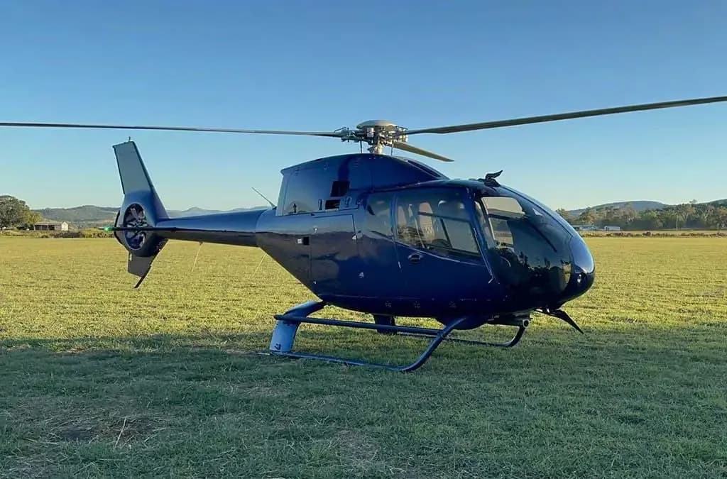 A dark blue helicopter with skids sits on a green grassy field under a clear blue sky.