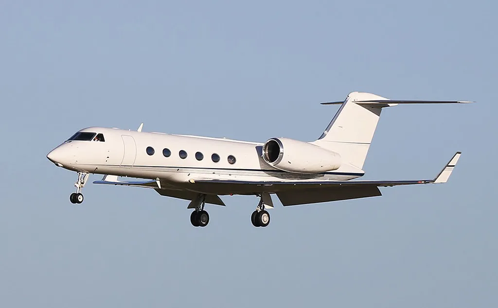 A white Gulfstream business jet with landing gear extended, flying against a clear sky.