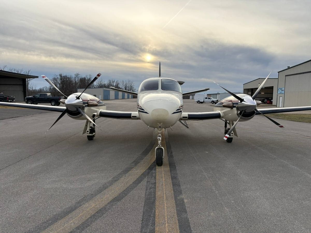 Front view of a white twin-engine propeller aircraft with dark stripes on an airport tarmac at sunset.