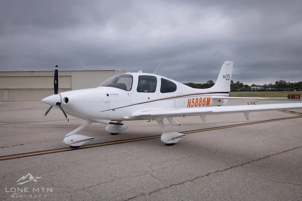 A white Cirrus SR20 aircraft with orange and black stripes, registration N5886M, parked on an airfield under a cloudy sky.