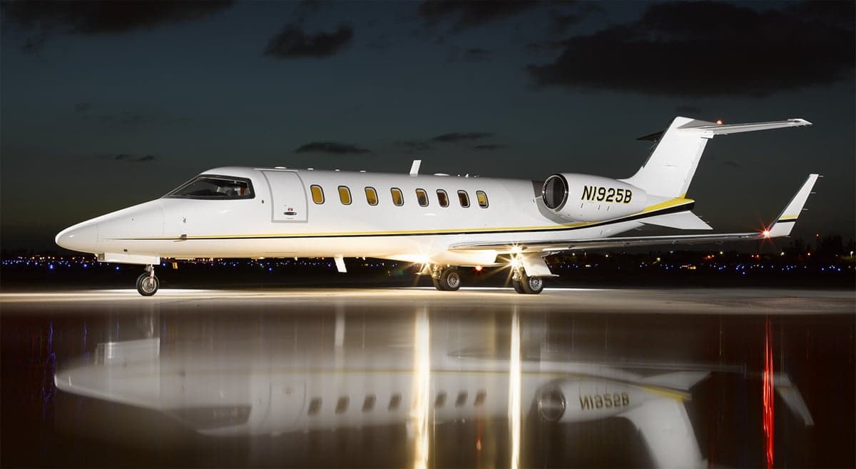 A white Learjet 60, registration N1925B, with yellow and black stripes, parked on a wet tarmac at night.