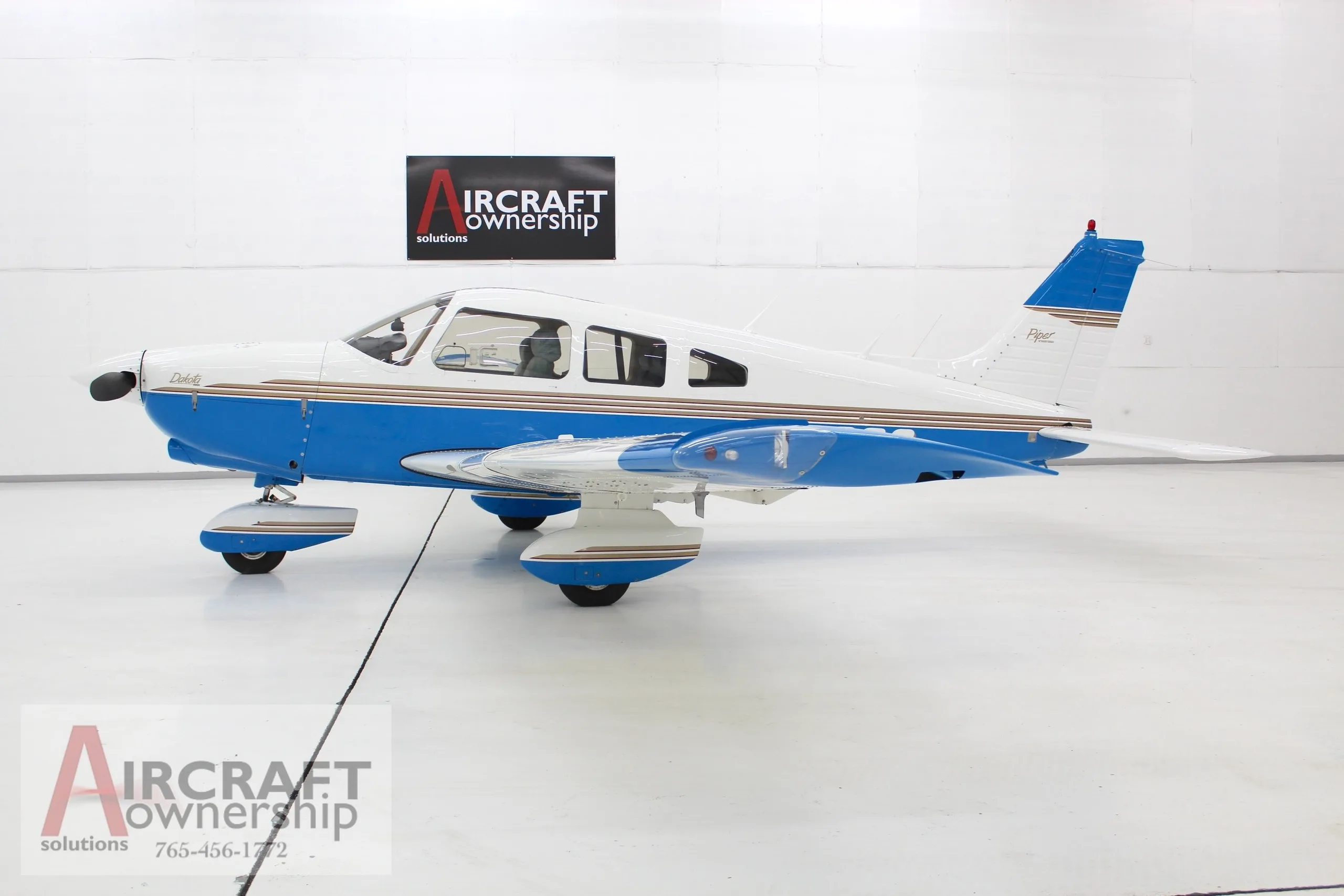 A white and blue Piper Dakota aircraft with gold stripes, viewed from the side in a hangar.