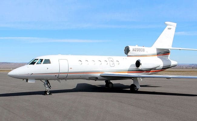 A white Dassault Falcon 900 business jet with grey and orange stripes is parked on an airport tarmac under a clear blue sky.