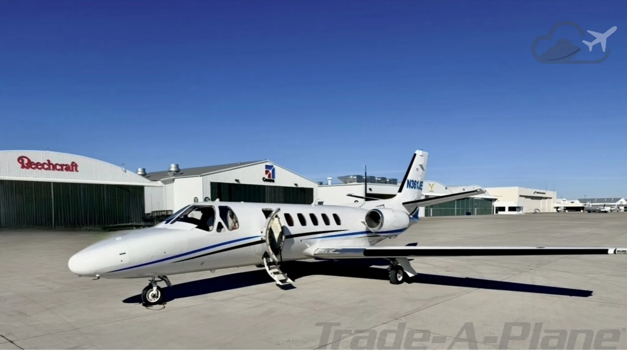 A white business jet with blue stripes and registration N361JE is parked on an airfield with its door open, hangars in the background.