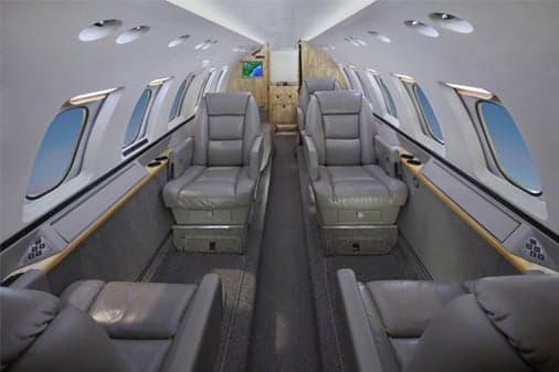 The interior cabin of a private jet with grey leather seats and large windows.