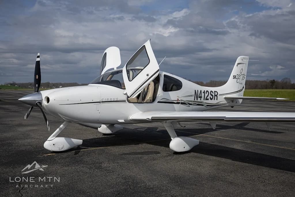 A white Cirrus SR22T aircraft with open doors is parked on an asphalt runway under a cloudy sky.