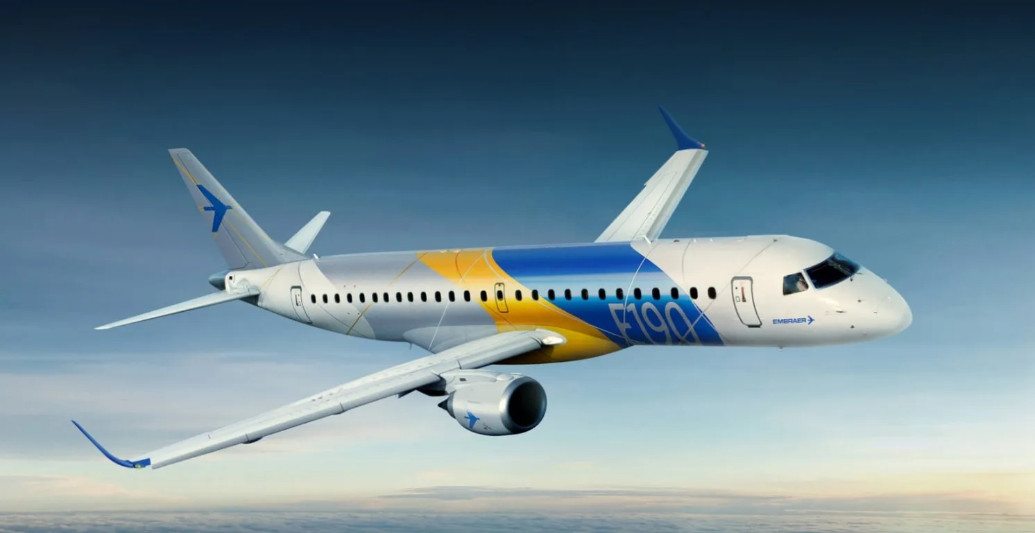 A white and blue Embraer E190 aircraft with yellow accents flies against a cloudy sky.