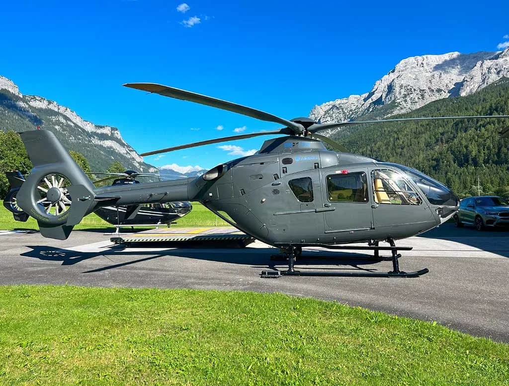 A grey Airbus H135 helicopter is parked on an asphalt pad with mountains in the background.