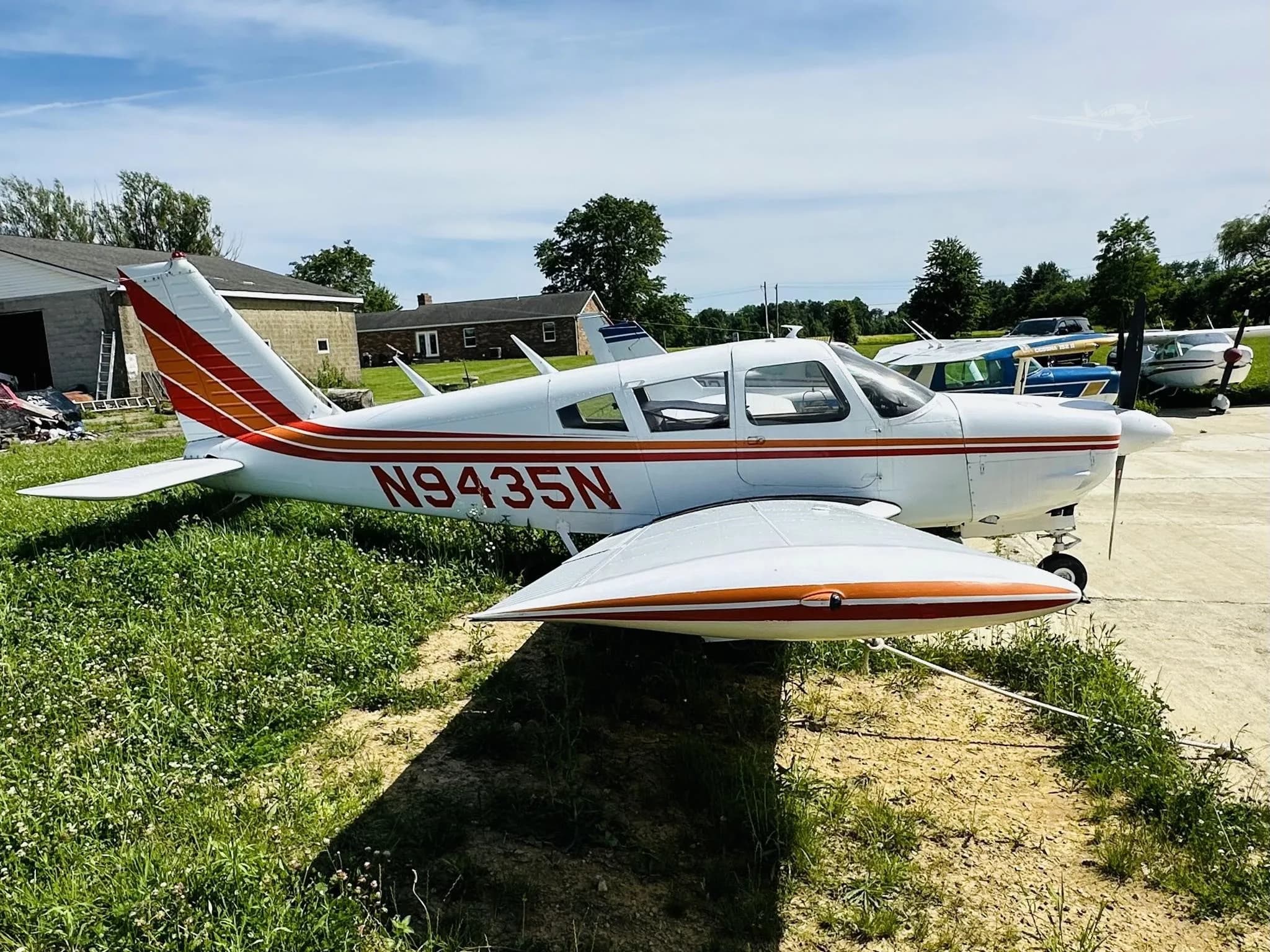 A white single-engine propeller aircraft with red and orange stripes, registration N9435N, parked on grass next to a concrete pad under a blue sky.