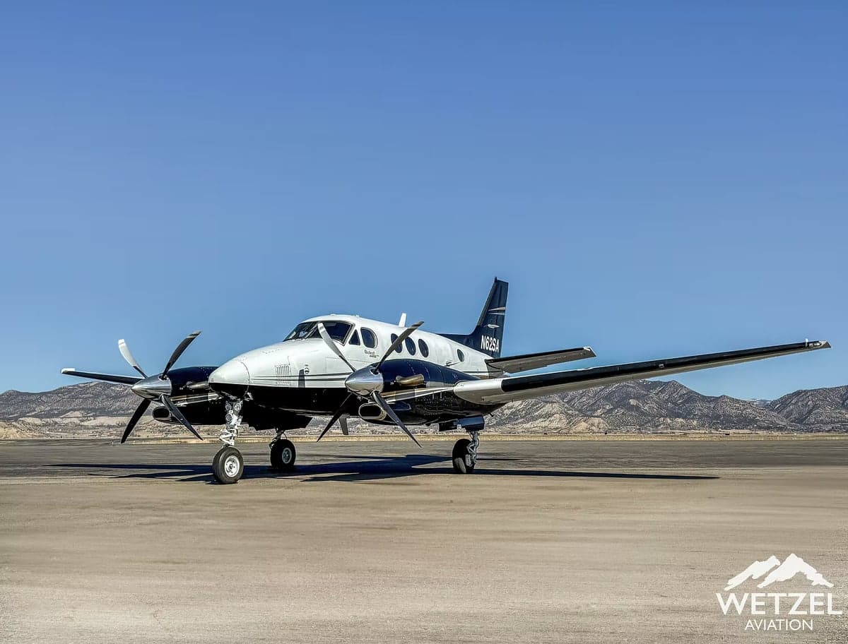 A black and white twin-engine turboprop aircraft, N62SA, is parked on an airfield with mountains in the background.