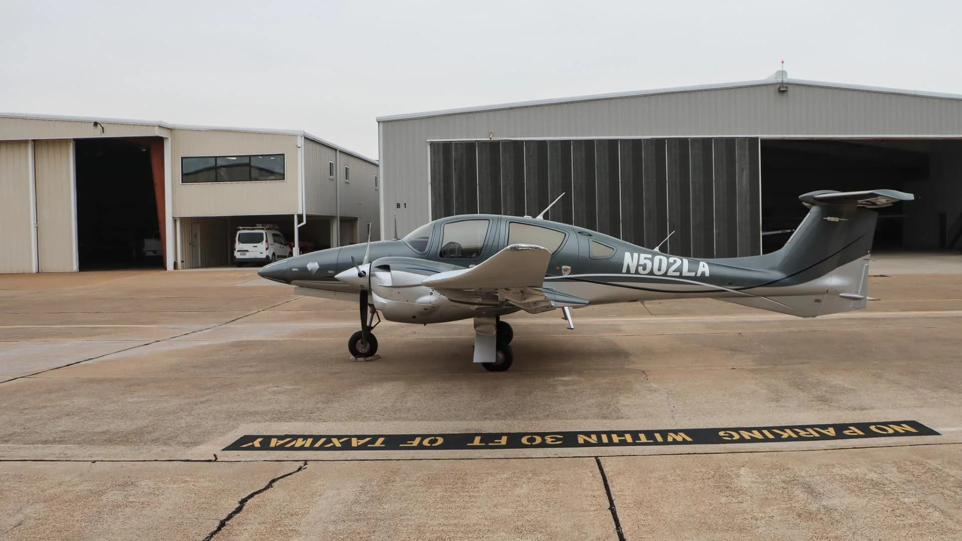 A gray and silver Diamond DA62 aircraft, N502LA, parked on an airport tarmac with hangars in the background.