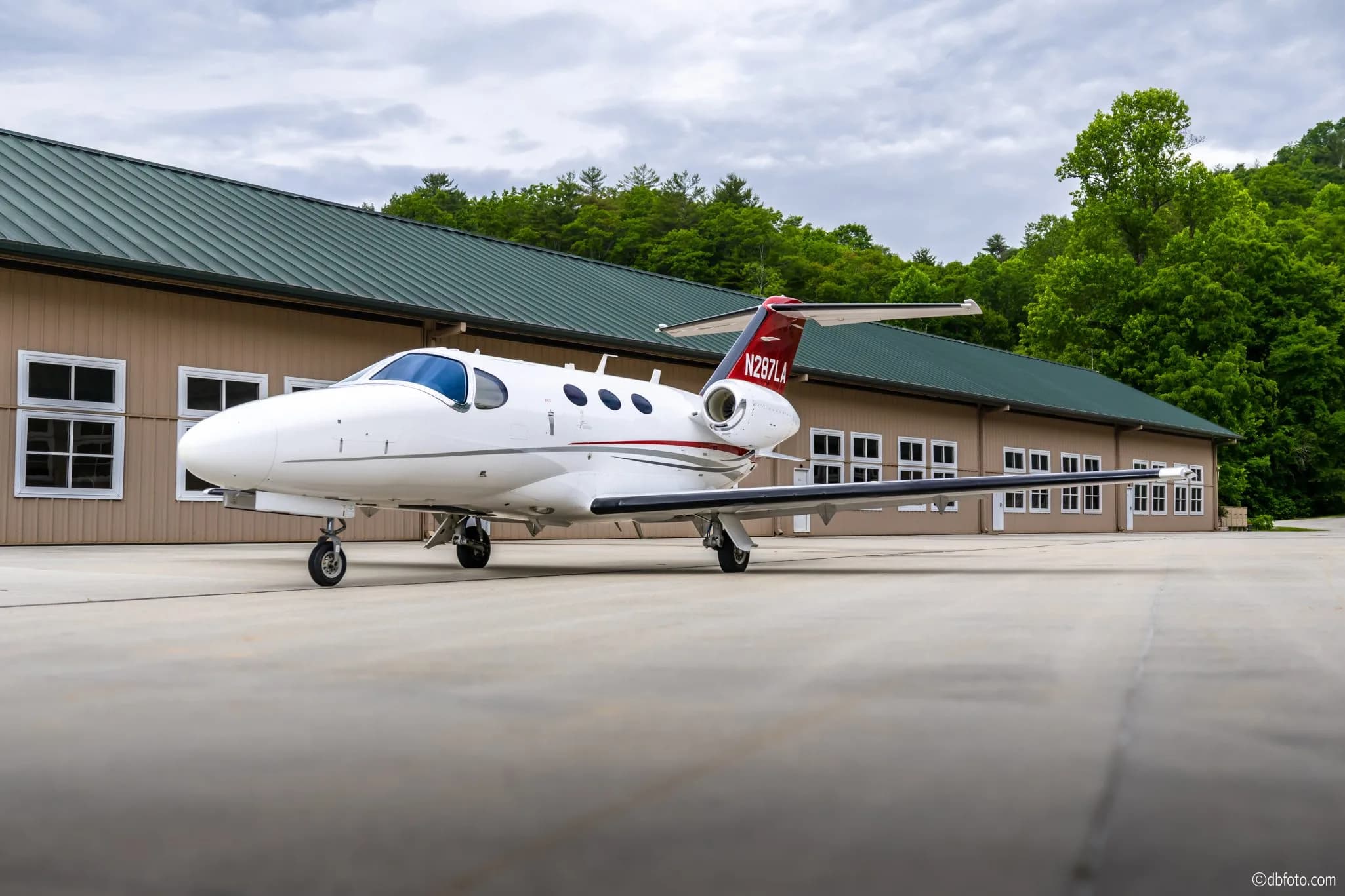A white Cessna Citation Mustang business jet with red and grey accents parked on an tarmac in front of a hangar.