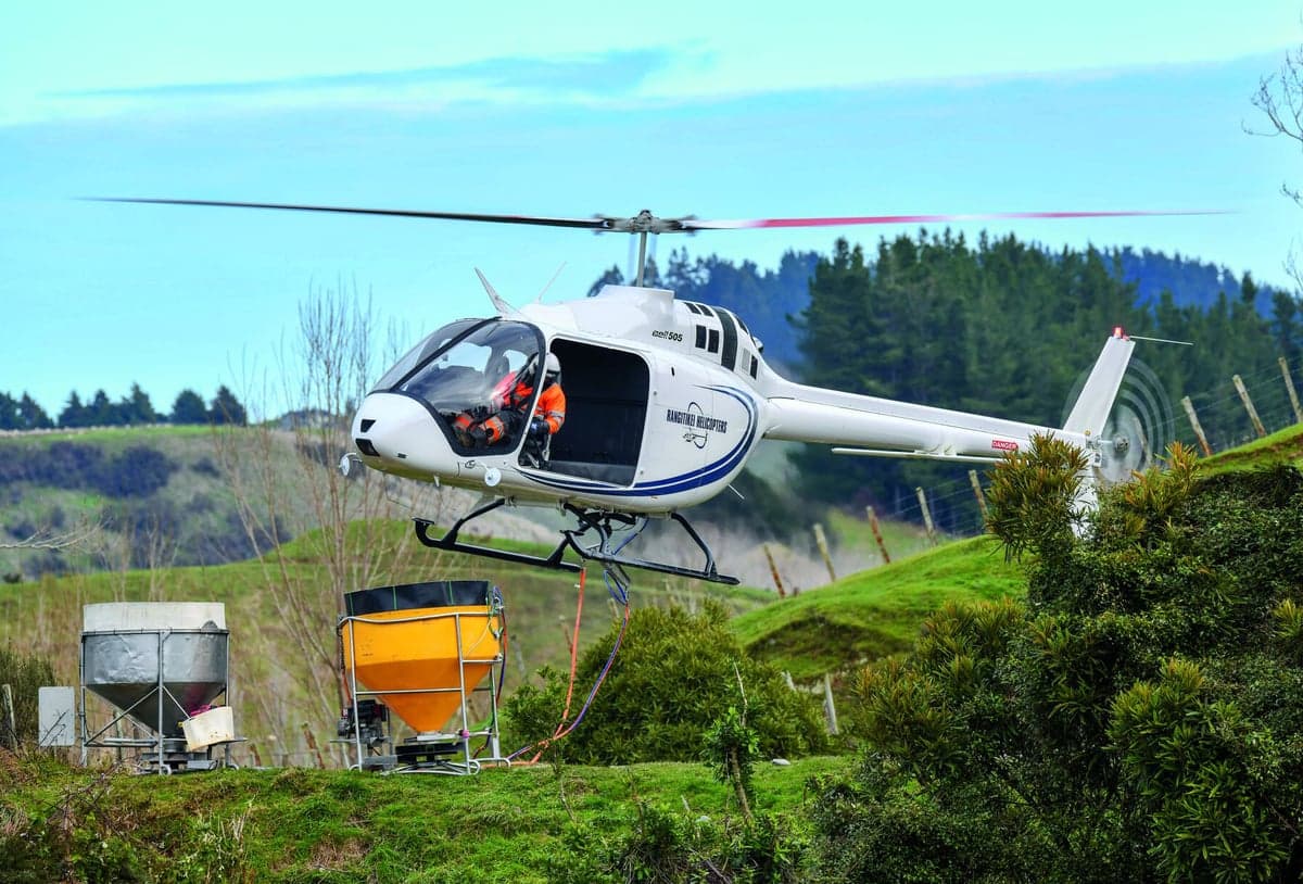 A white and blue Bell 505 helicopter in flight, with a person visible in the cockpit.