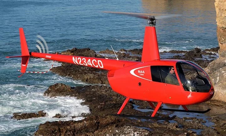 A red Robinson R44 helicopter, registration N234CD, lands on a rocky shore.