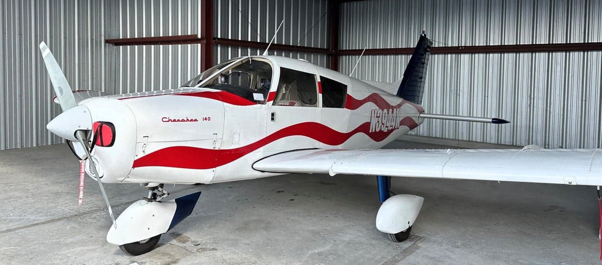 A Piper Cherokee 140 aircraft with a red, white, and blue paint scheme is parked in a hangar.