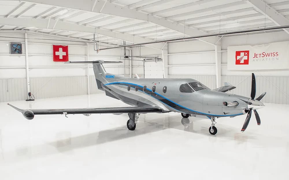 A silver Pilatus PC-12 turboprop aircraft with blue and grey stripes is parked inside a white hangar with Swiss flags and JetSwiss Aviation branding.