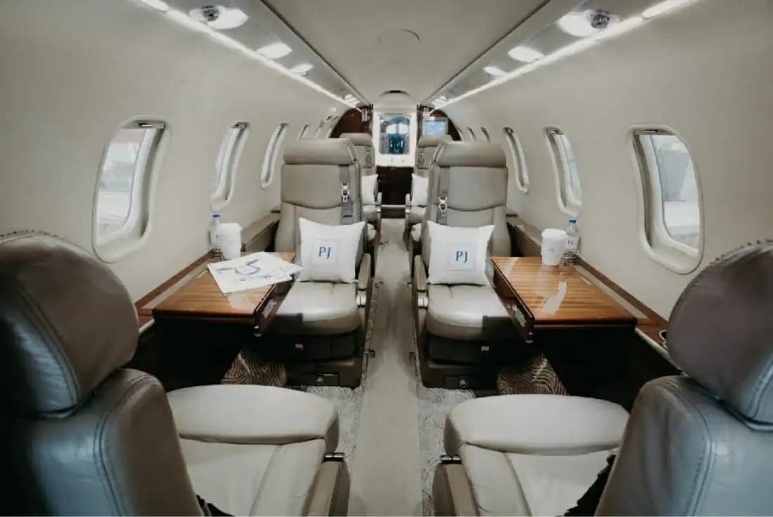 View of a luxurious aircraft cabin with light-colored leather seats, wooden tables, and white pillows featuring 'PJ' initials.