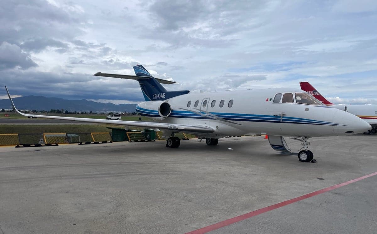 A white business jet with blue stripes (XB-OAE) parked on a tarmac under a cloudy sky, with mountains in the background.