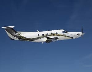 A white Pilatus PC-12 aircraft with black and gold swooping livery flies against a clear blue sky.