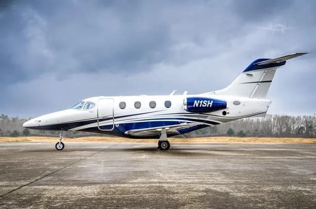 A white and blue business jet, tail number N1SH, parked on an airfield under a cloudy sky.