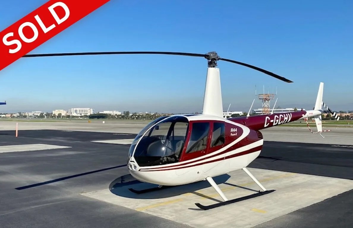 A maroon and white Robinson R44 Raven II helicopter is parked on an airfield under a clear blue sky.