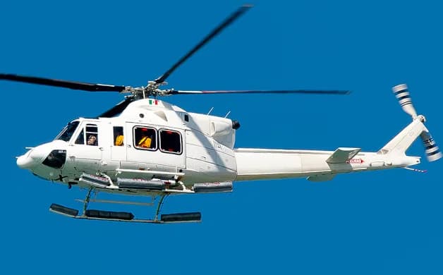 A white Agusta-Bell AB 412 helicopter with an Italian flag on its tail flies against a clear blue sky.