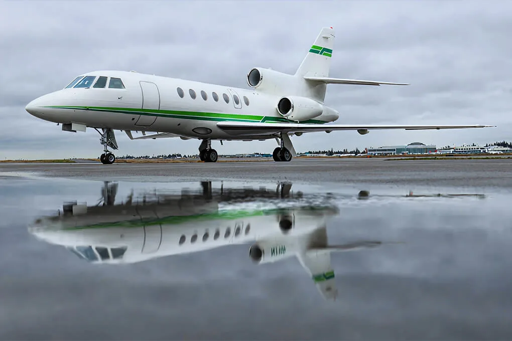 A white Dassault Falcon business jet with green stripes on a wet tarmac, reflecting in puddles.