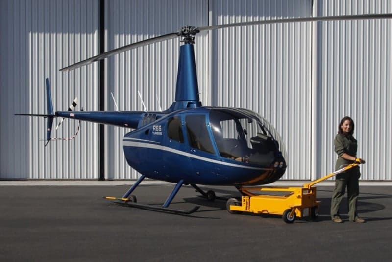 A blue Robinson R66 helicopter with a woman and a yellow tug in front of a hangar.