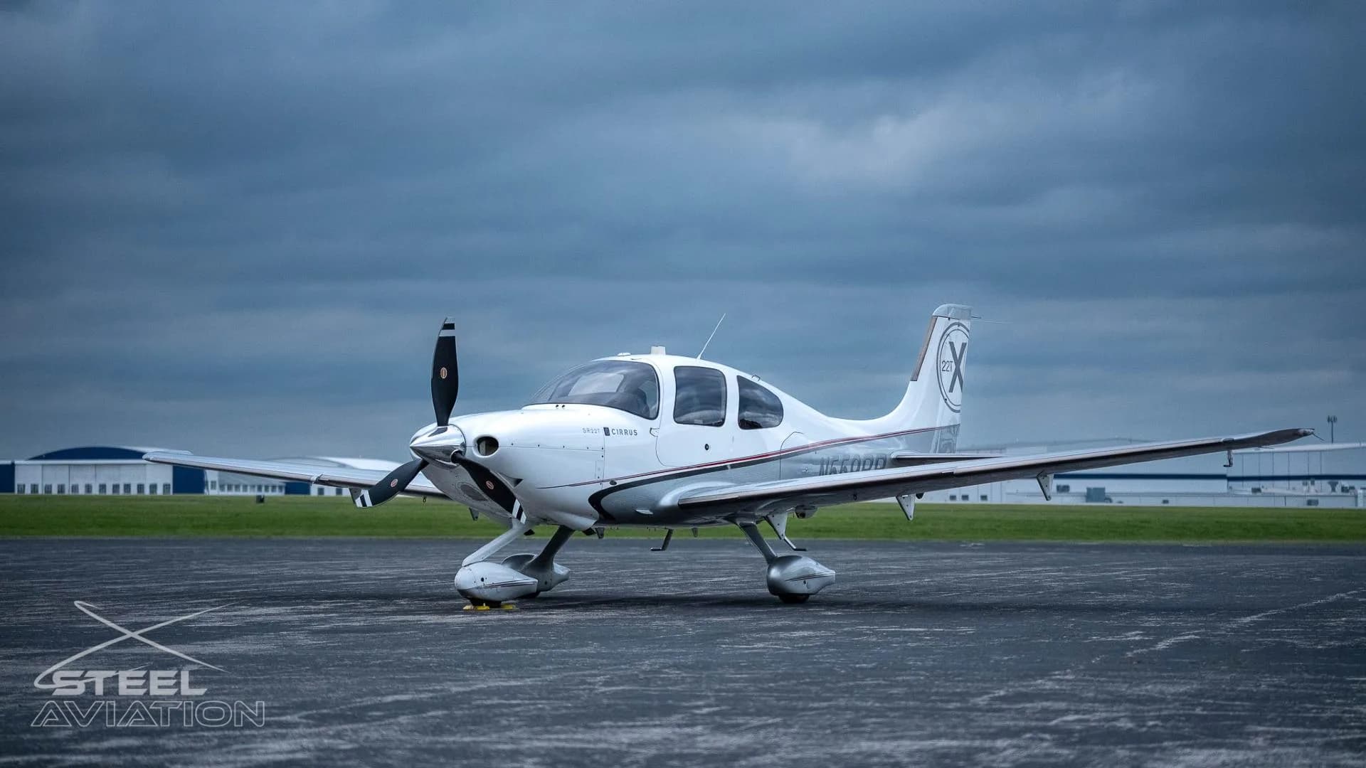 A white and silver Cirrus SR22T aircraft with a three-bladed propeller sits on an airfield under a cloudy sky.