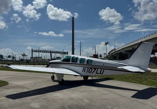 A white single-engine aircraft, N107LU, with a black stripe, parked on a tarmac with a highway overpass in the background.