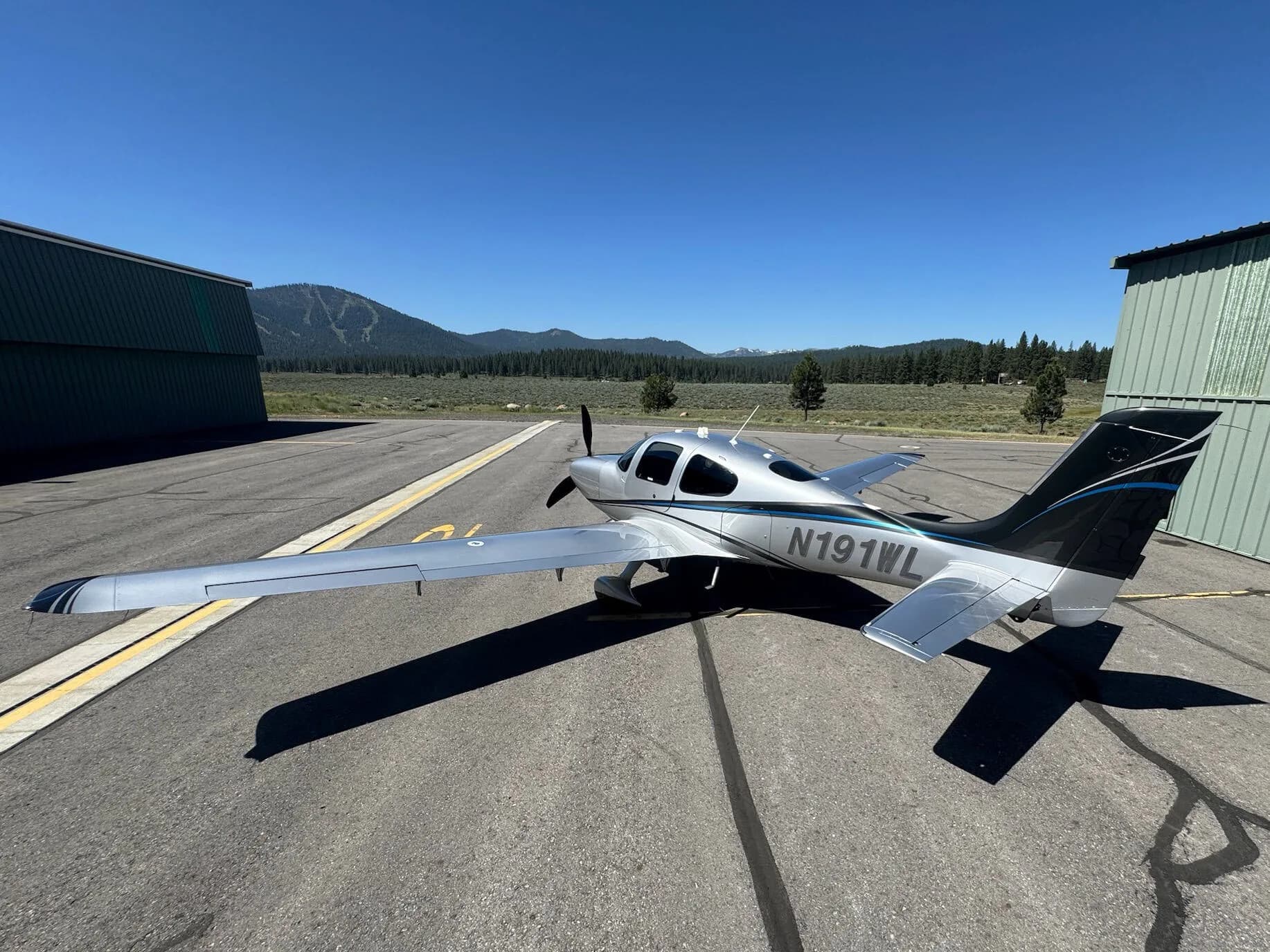 A silver and black Cirrus SR22T aircraft with N191WL on the tail, parked on an asphalt apron with hangars and mountains in the background.