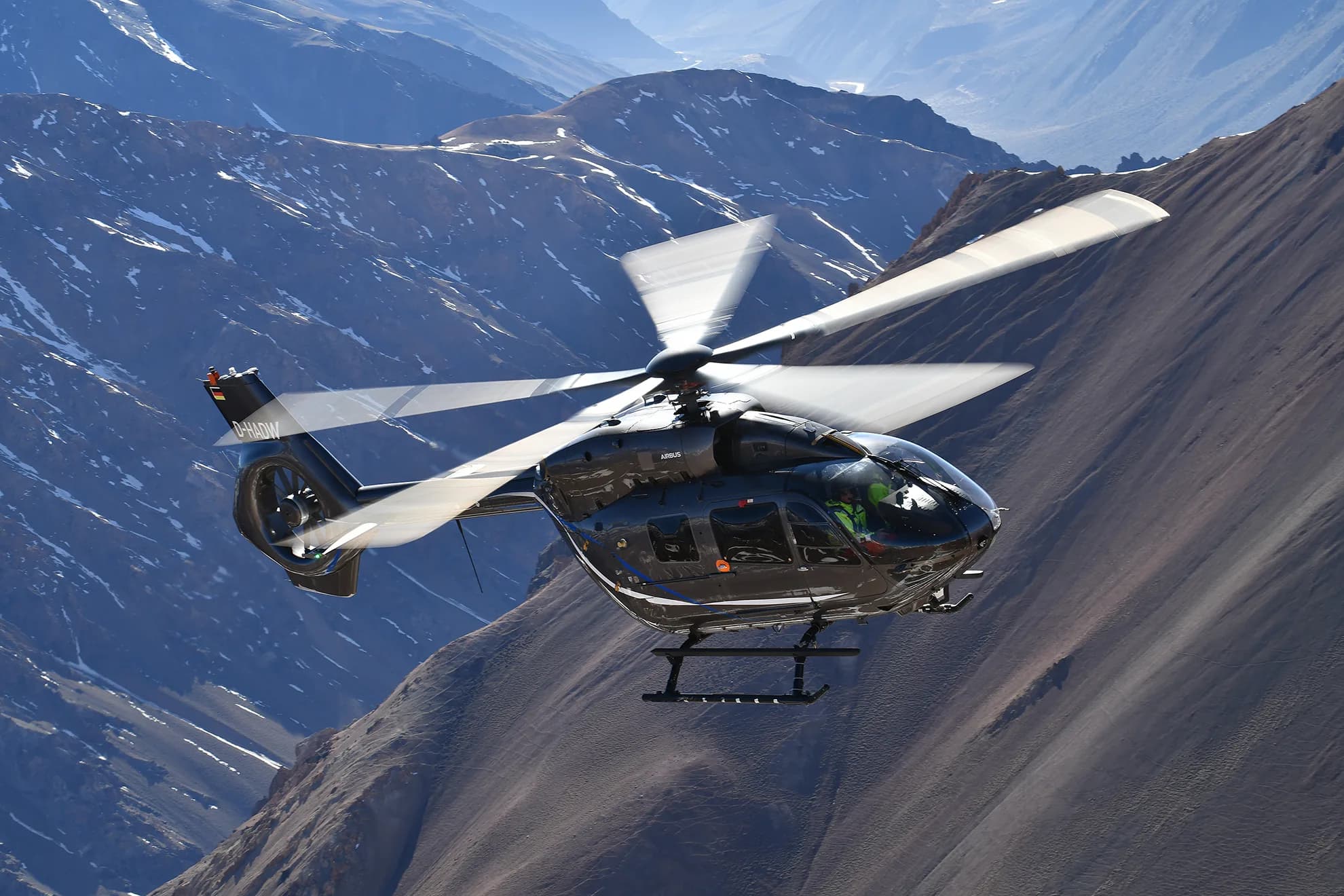 A black Airbus H145 helicopter with registration D-HAUW flying over a mountainous landscape.