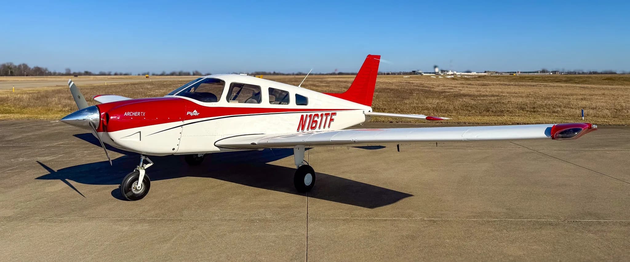 A red and white Piper Archer TX aircraft, tail number N161TF, parked on an asphalt tarmac under a clear blue sky.