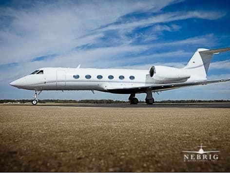 A white Gulfstream G550 business jet is parked on an airfield under a blue sky with clouds.