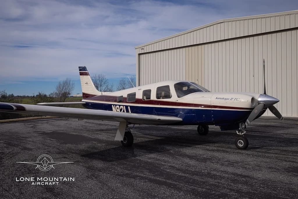 A Piper Saratoga II TC aircraft (N92LL) with blue and white paint scheme parked on an asphalt apron in front of a hangar.