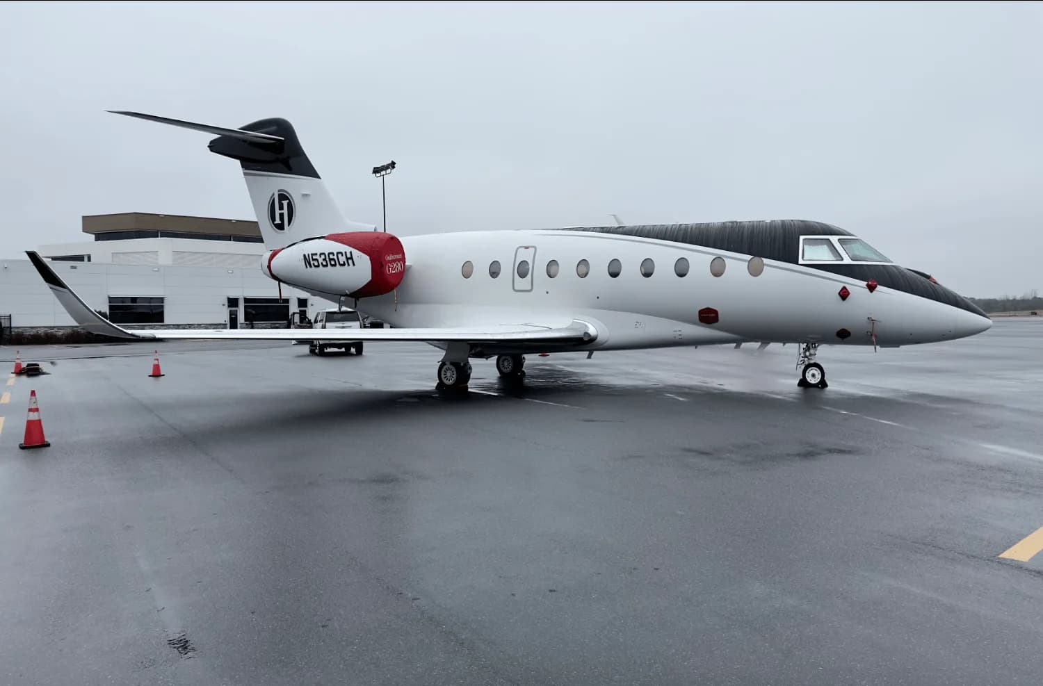 A white and black Gulfstream G280 business jet with red accents on the tail and engine nacelle, parked on a wet tarmac.