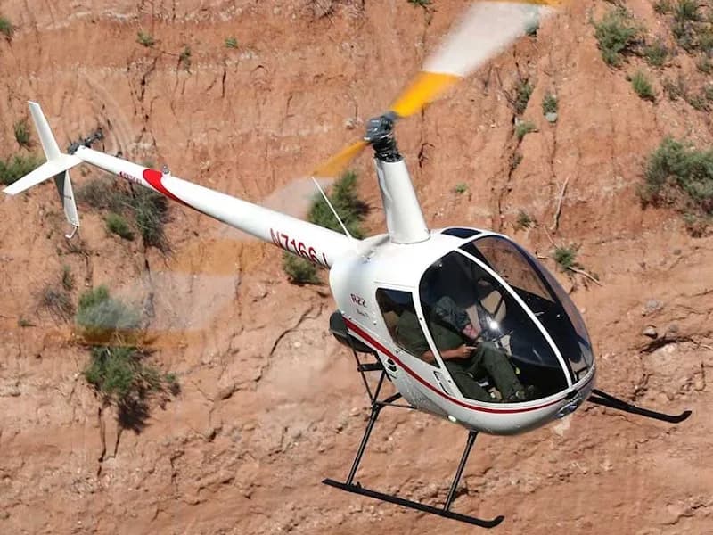 A white and red Robinson R22 helicopter in flight near a canyon wall.