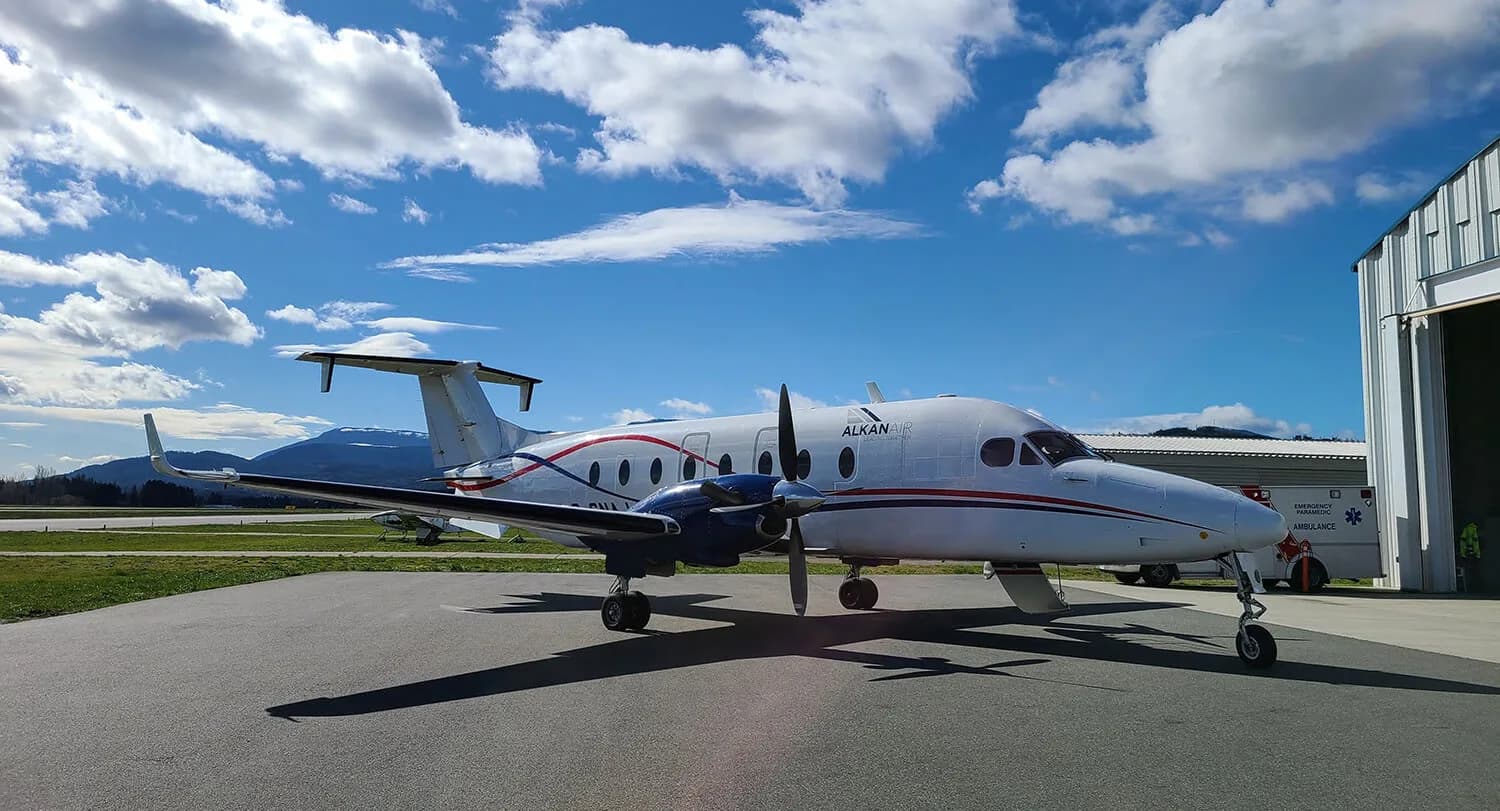 A white and blue Alkan Air twin-turboprop aircraft with red stripes is parked on an airfield next to a hangar under a blue sky.