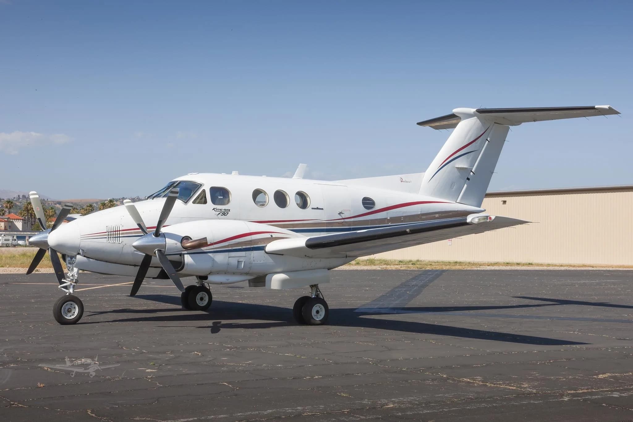 A white King Air F90 with red and blue stripes is parked on an airport tarmac under a clear sky.