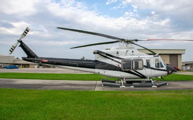 A white and black helicopter with floats on its landing gear is parked on an asphalt pad next to green grass under a partly cloudy sky.