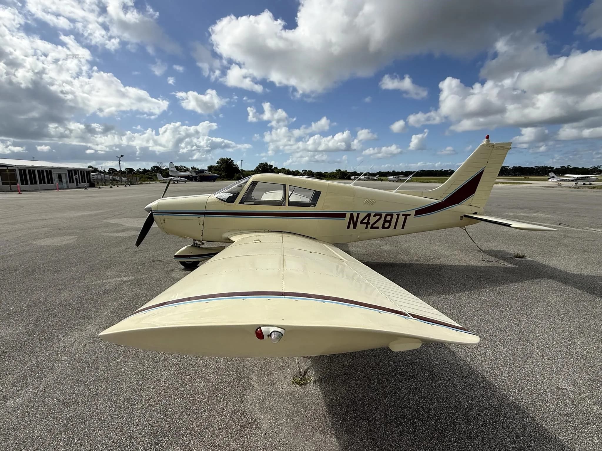A cream-colored single-engine propeller aircraft with maroon and blue stripes, registration N4281T, parked on an asphalt tarmac under a partly cloudy sky.