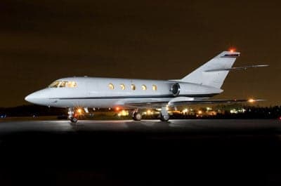A white business jet, likely a Dassault Falcon, is parked on a tarmac at night with its lights illuminated and city lights in the background.