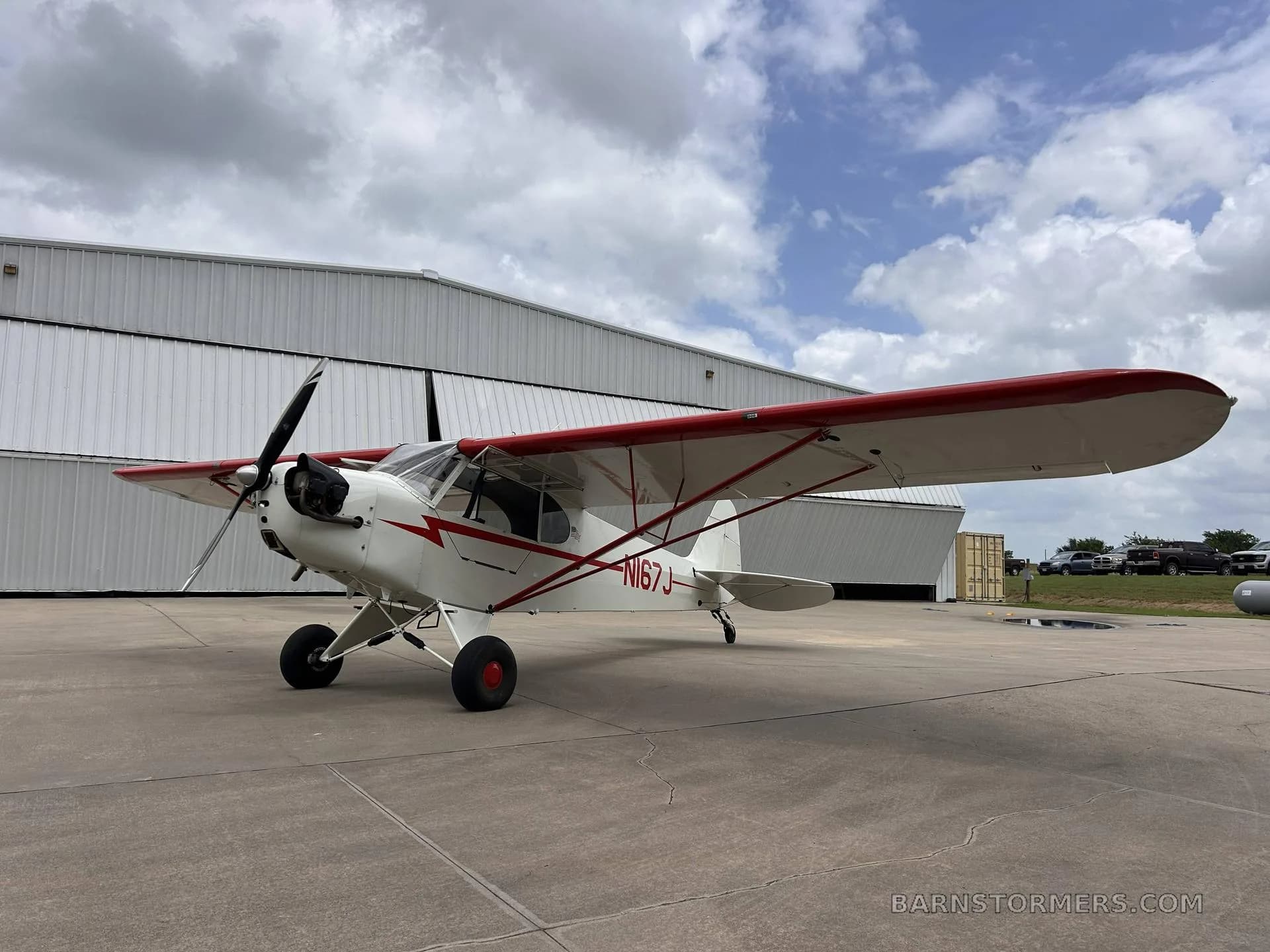 A white and red single-engine aircraft (N167J) with high wings is parked on an asphalt apron in front of hangars under a cloudy sky.