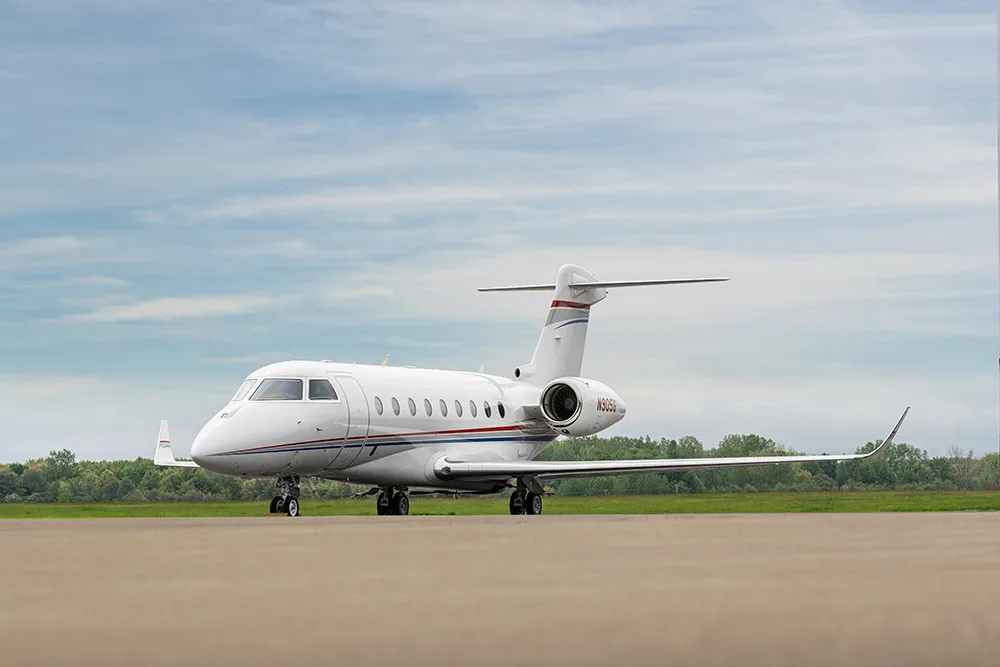 A white business jet with red and blue stripes on the tail and fuselage, parked on an airfield under a partly cloudy sky.