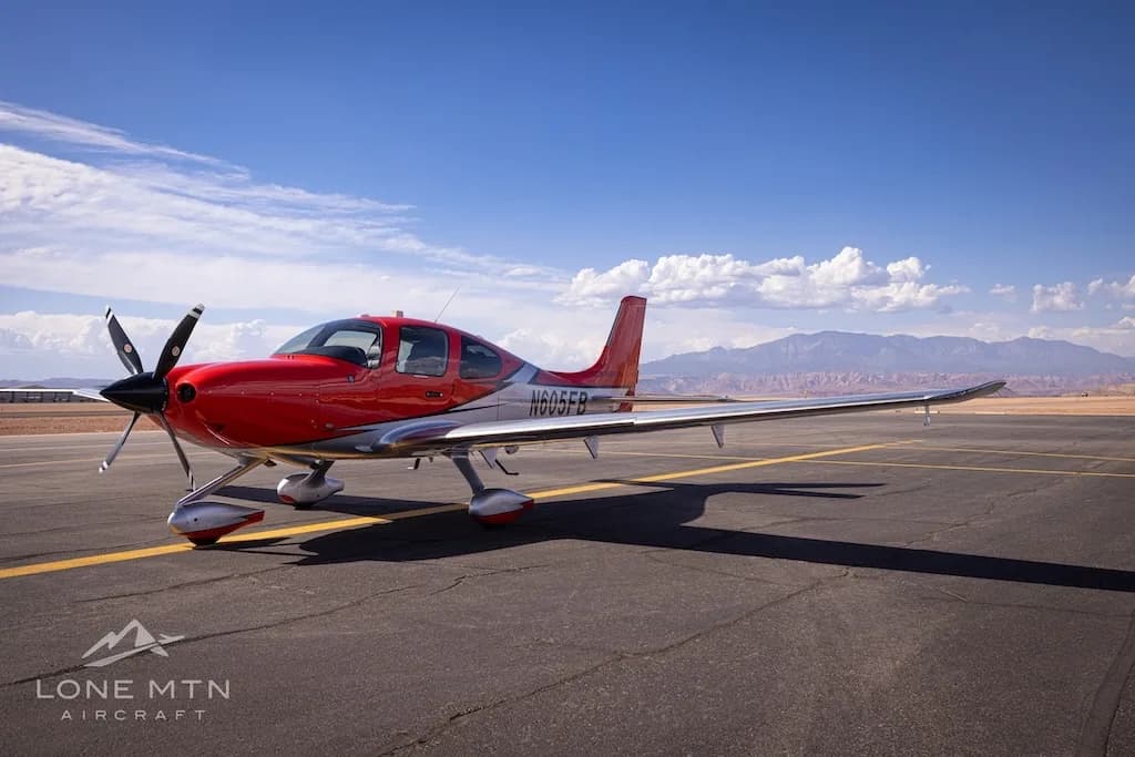 A red and silver Cirrus SR22T aircraft (N605FB) parked on an airfield under a blue sky with mountains in the background.