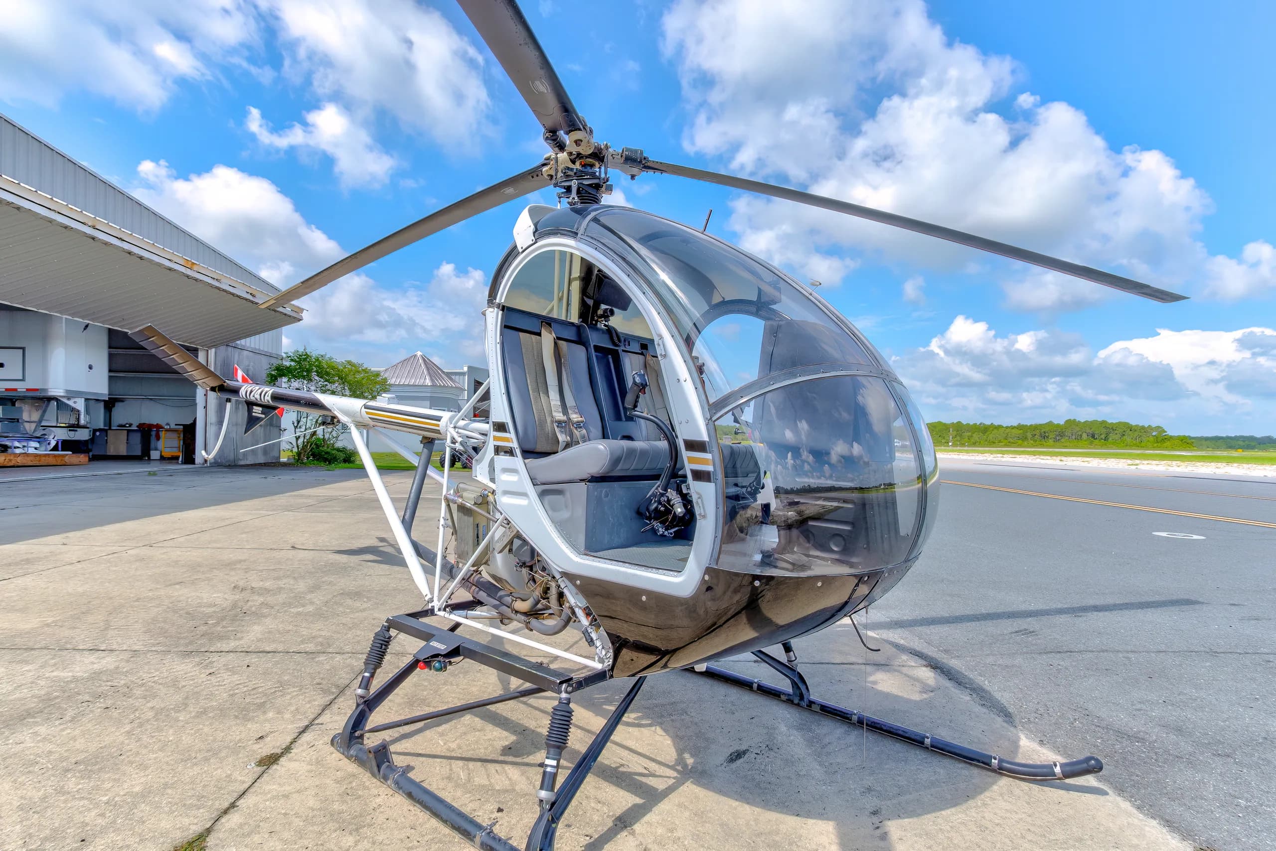 A black and white Schweizer 300CBi helicopter, N124TA, parked on an airfield tarmac next to an open hangar under a blue sky.