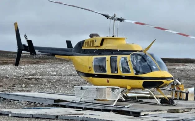 A yellow and black Bell 206 JetRanger helicopter with white and red main rotor blades sits on a rocky ground next to a wooden platform.