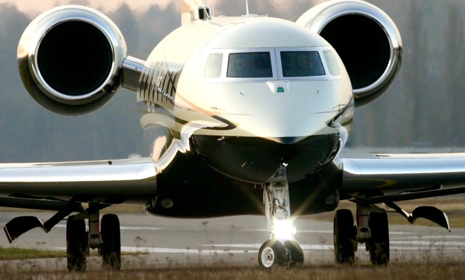 A front view of a white and black business jet with landing lights on, taxiing on a runway at dawn or dusk.