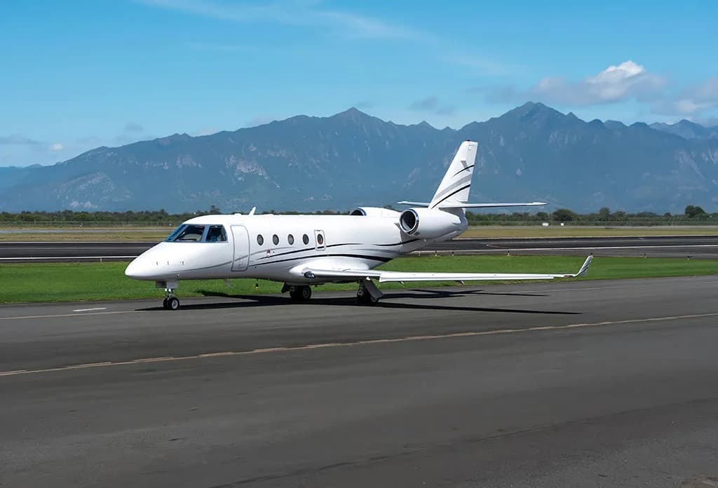 A white business jet with black and grey stripes is parked on a runway with mountains in the background.